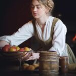 Woman in period clothing holding a bowl of apples in a rustic kitchen