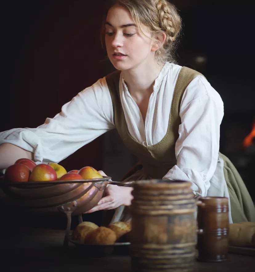 Woman in period clothing holding a bowl of apples in a rustic kitchen