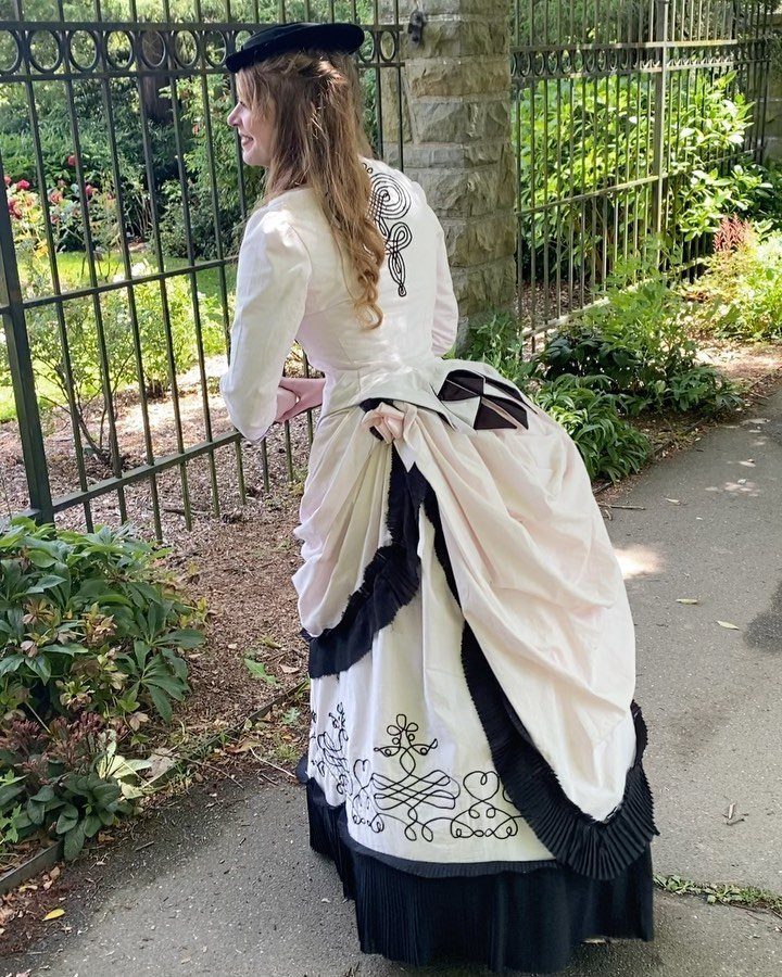 Woman in a cream Victorian dress with a large bustle standing by a fence