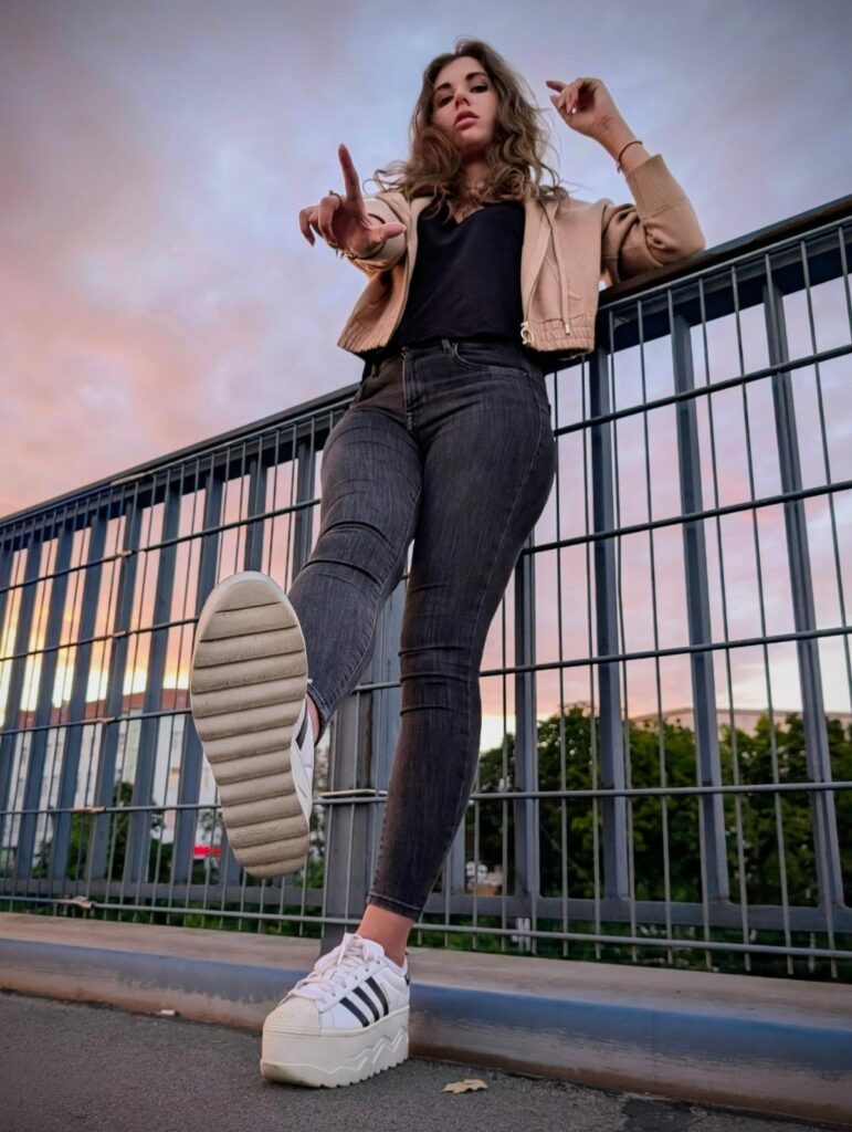 Woman in grey jeans and white platform Adidas sneakers posing by a railing