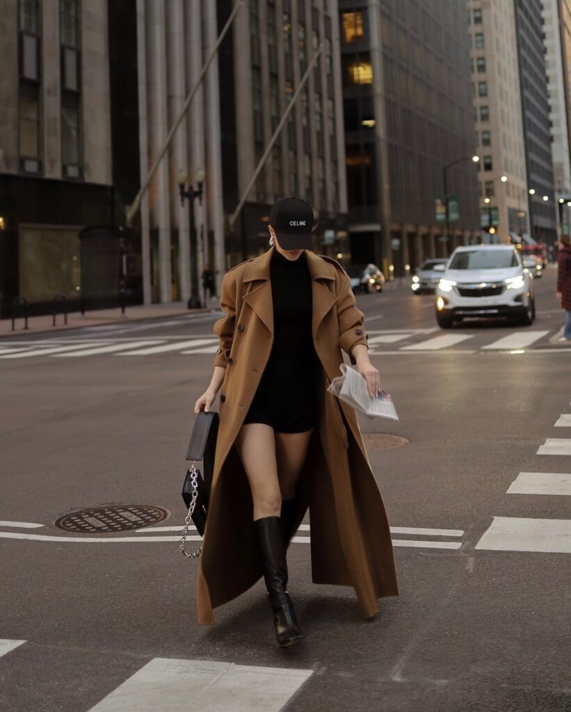Person in tan trench coat and black dress wearing black leather boots.