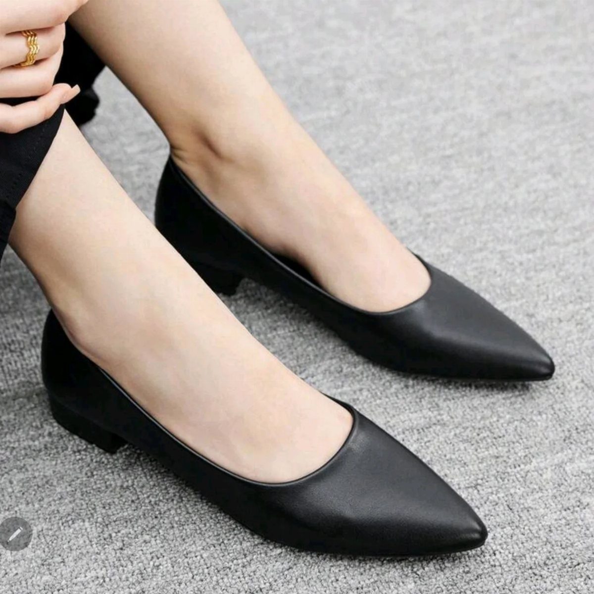 Black pointed-toe low heel pumps worn on feet standing on grey carpet.