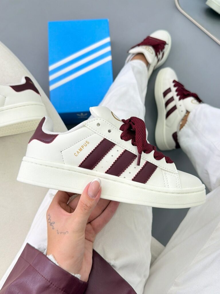 White Adidas Campus sneakers with burgundy stripes and accents held by a person.