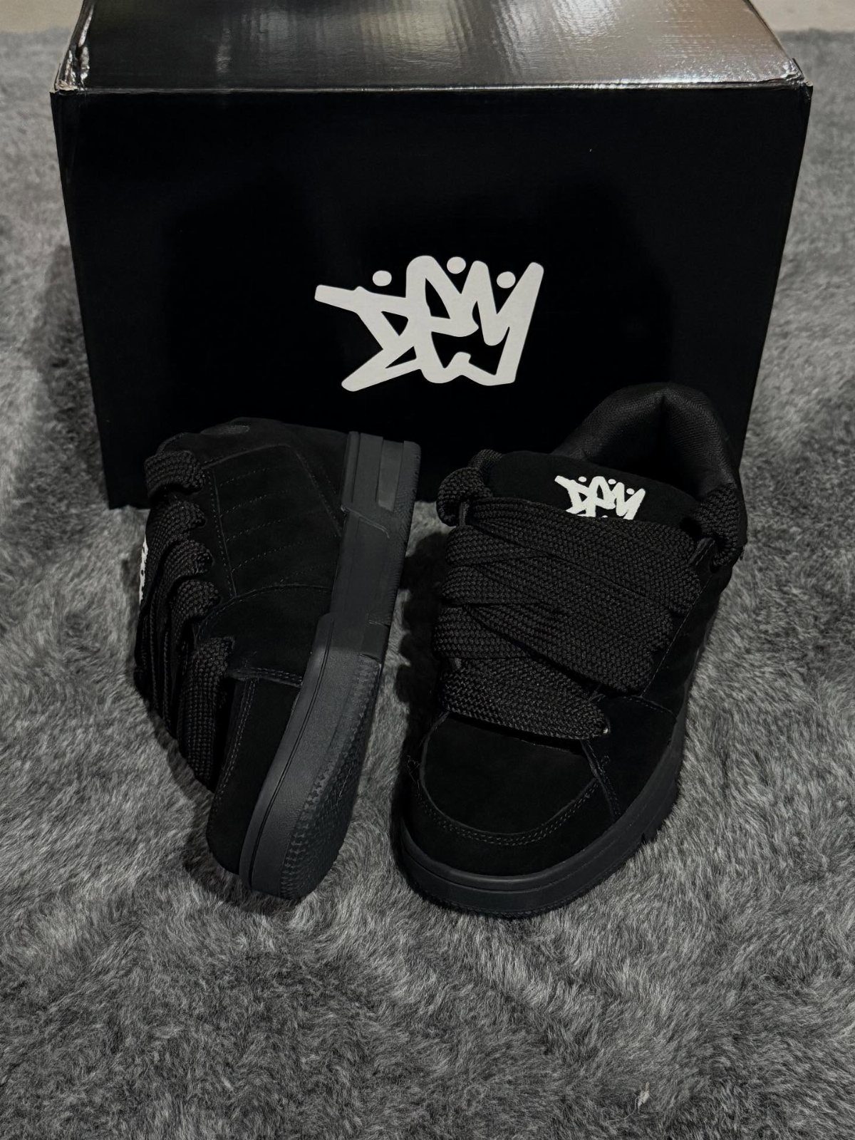 Black suede sneakers with thick chunky laces in front of a black box.