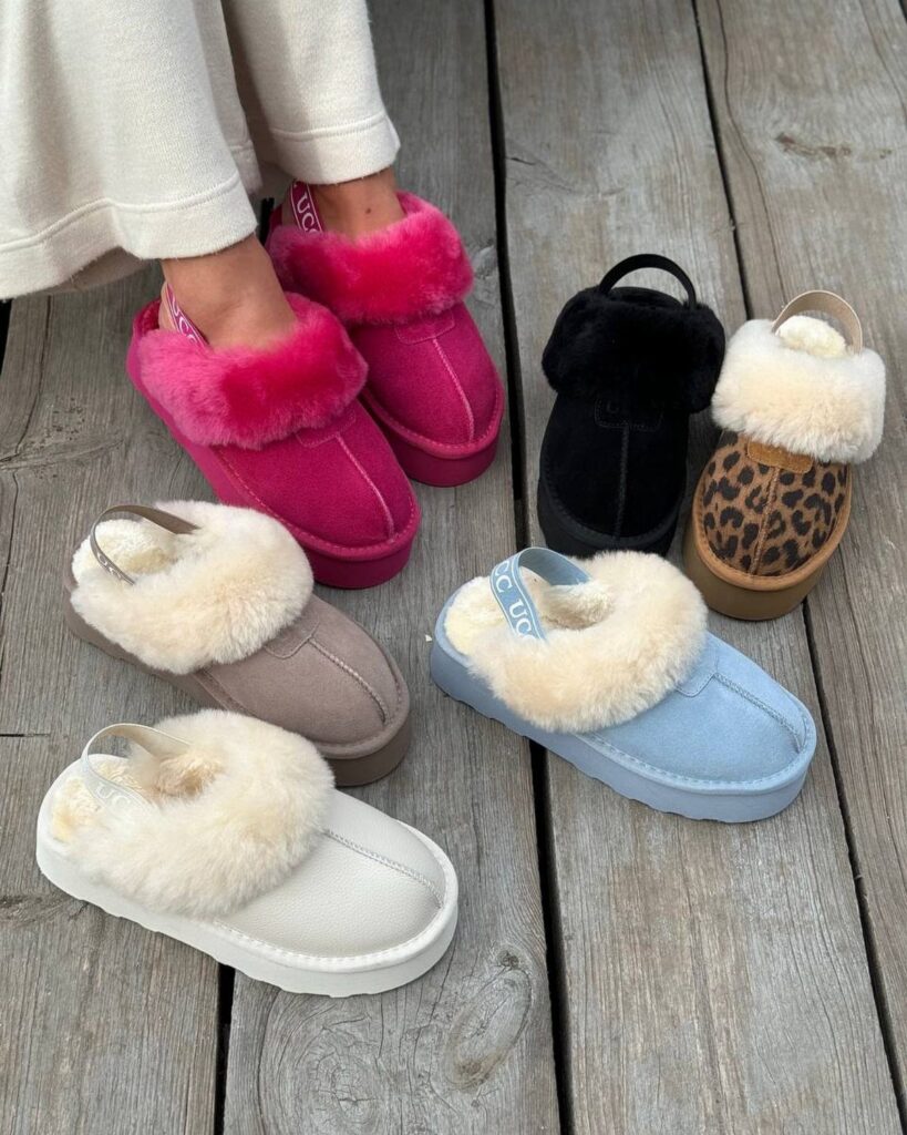 Various colorful sheepskin-lined Tasman slippers arranged on a weathered grey wooden deck.