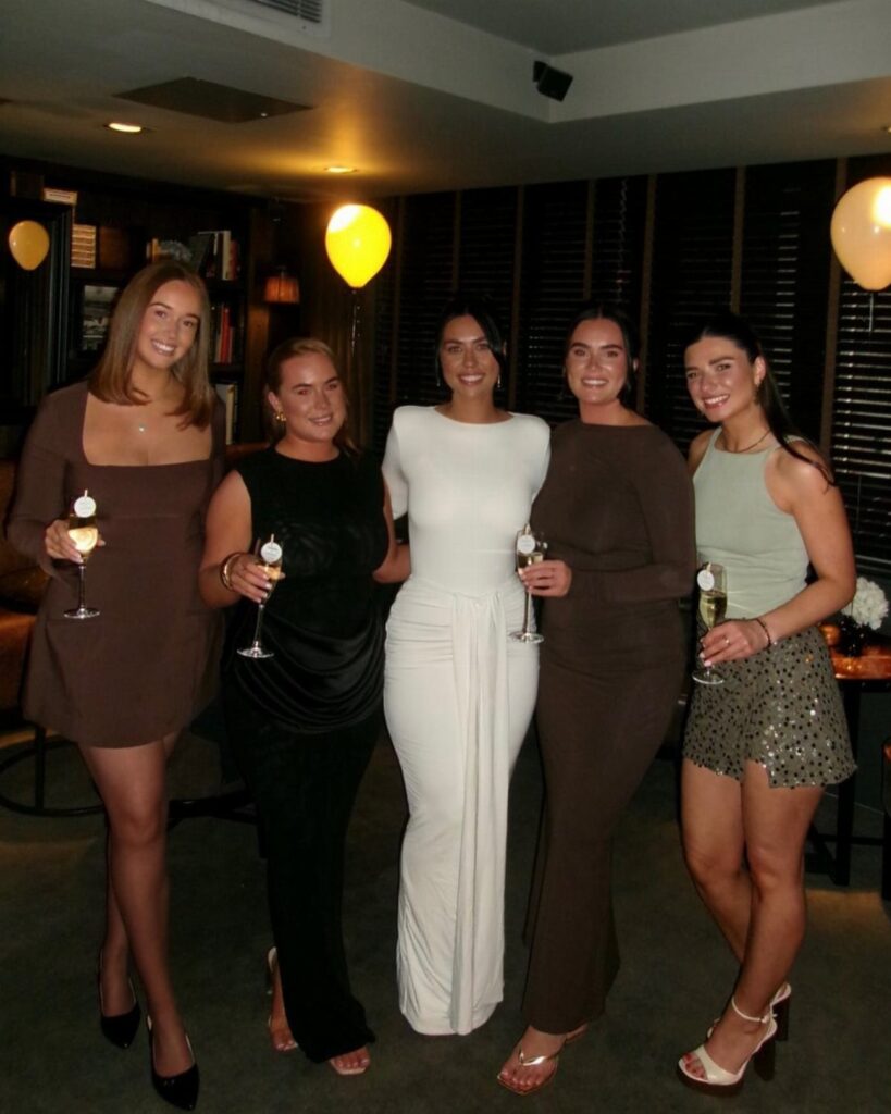 Five women in cocktail dresses and evening wear posing with champagne glasses indoors.