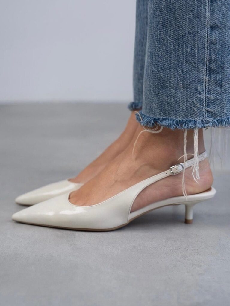 White pointed-toe kitten heels with slingback straps worn with blue frayed denim jeans.