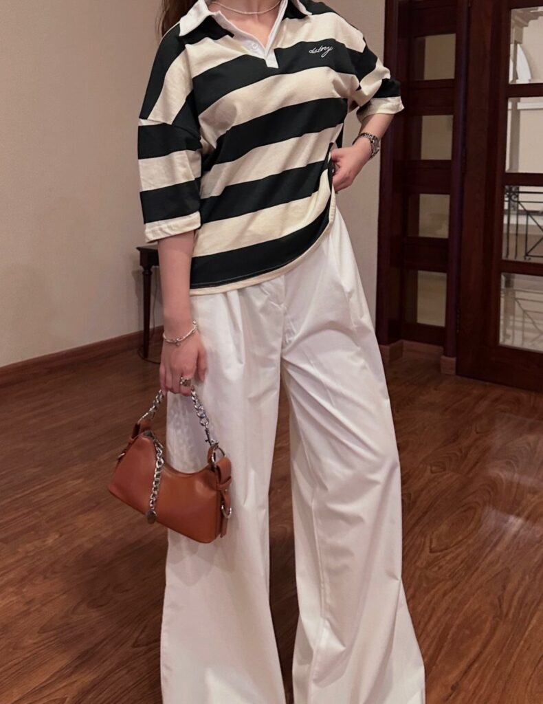 Person wearing a striped polo shirt and wide-leg white trousers holding a brown handbag.