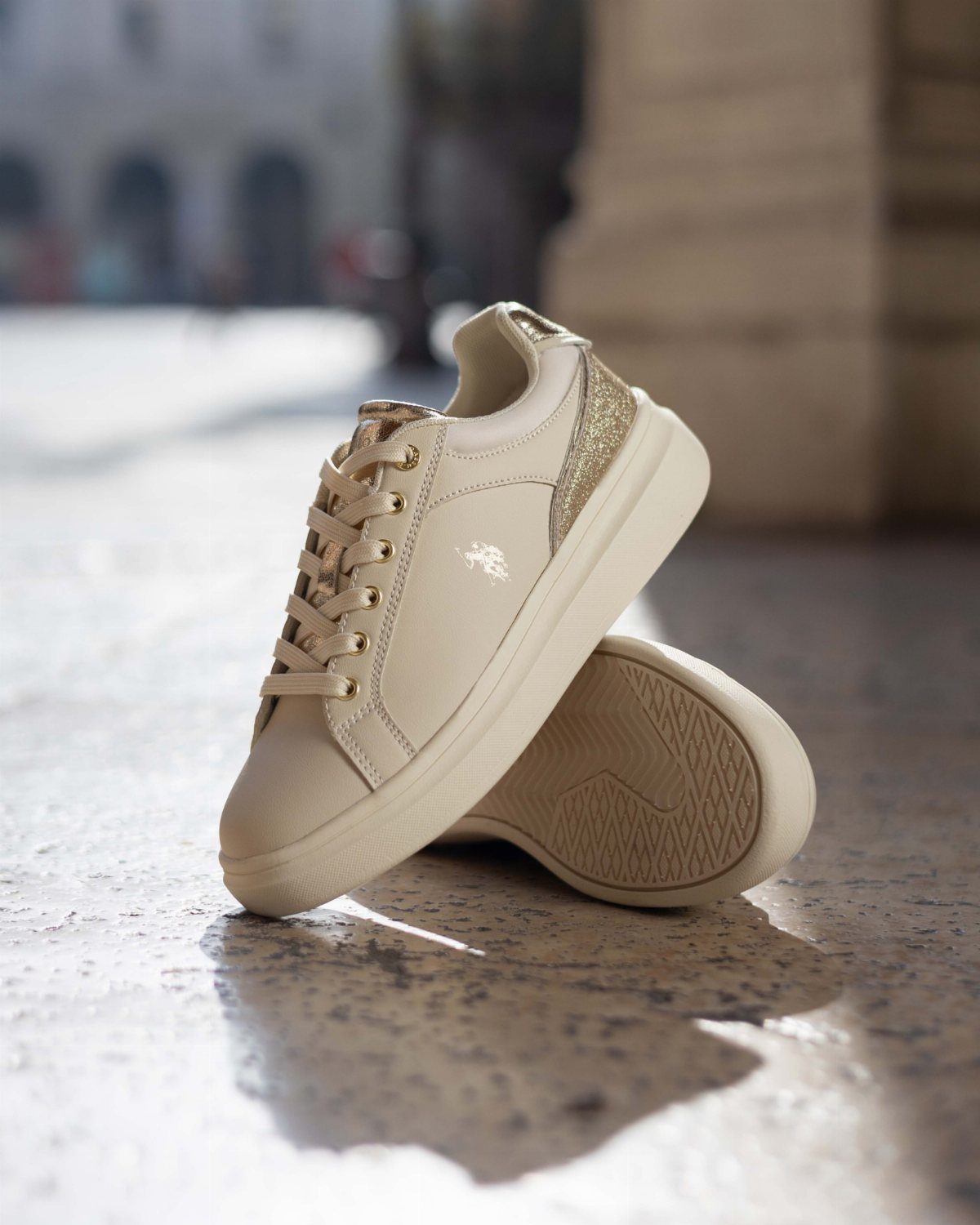 Beige women's sneakers with gold glitter details resting on a stone pavement.