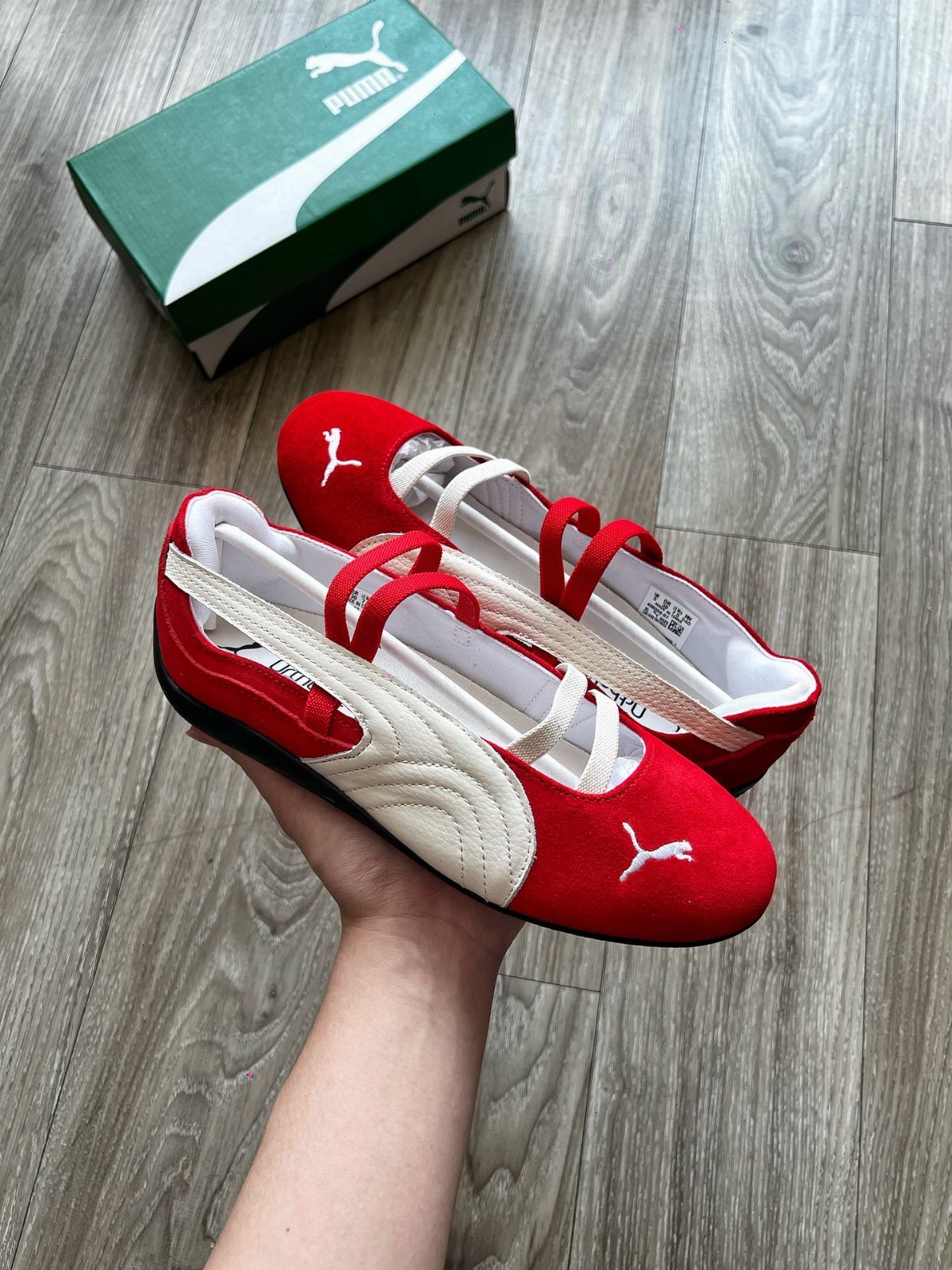 Red and white Puma ballet-style sneakers held over a grey wooden floor.