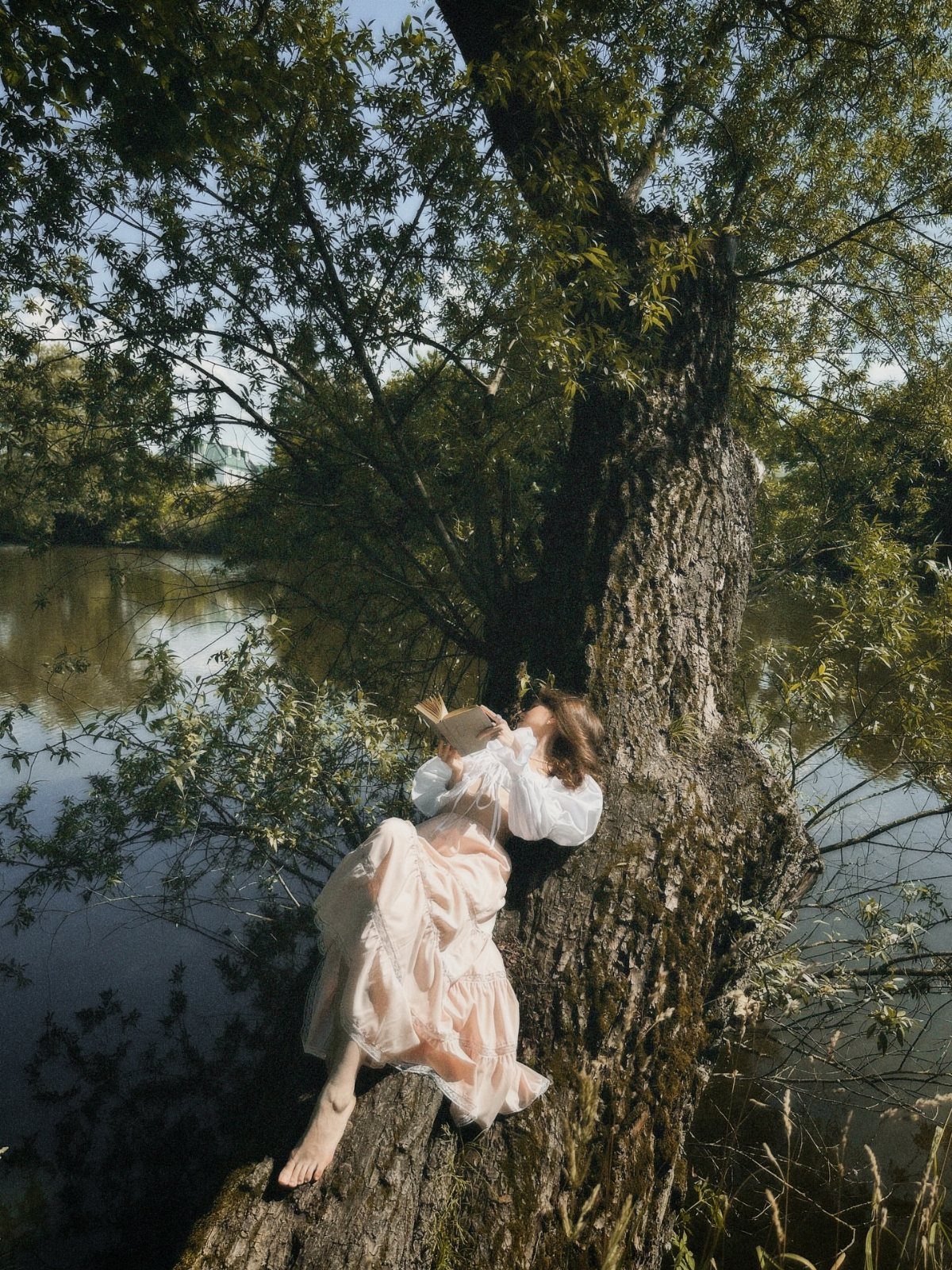 Woman in a flowing white dress leaning against a tree by a river