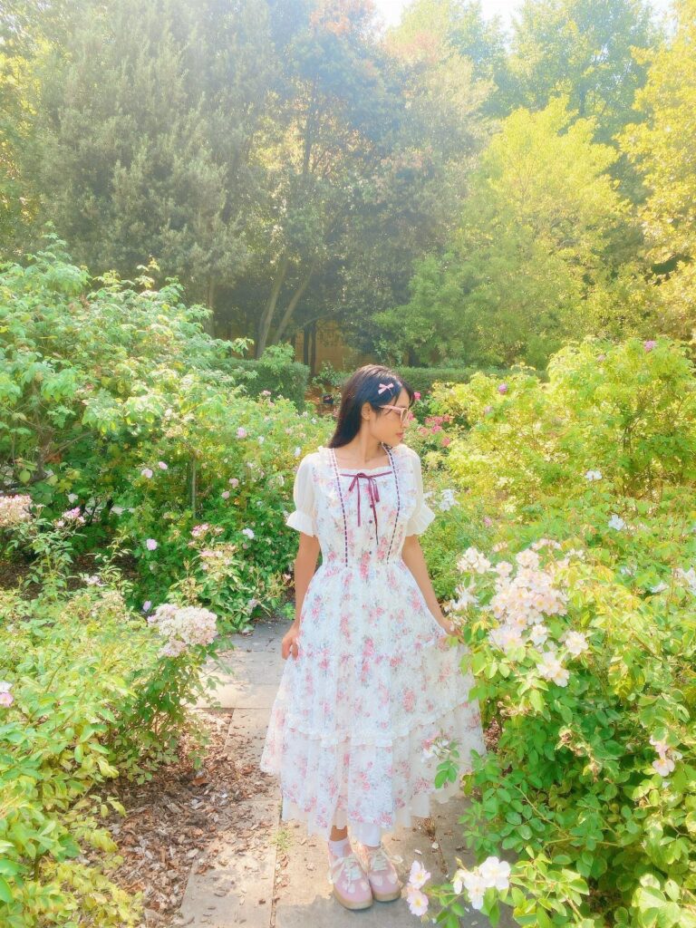 Woman in a white floral dress standing among white roses in a garden.
