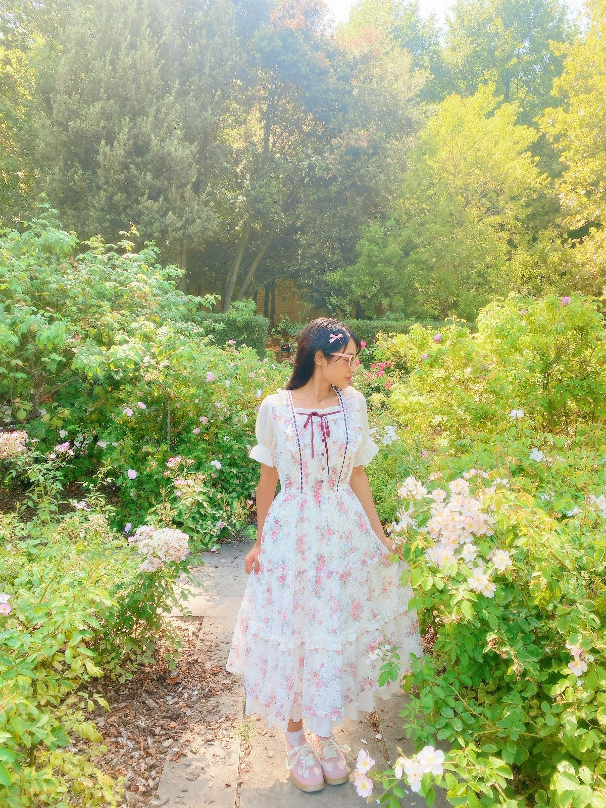 Woman in a white floral dress standing among white roses in a garden.