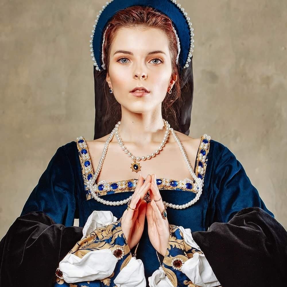 Woman in a blue velvet Tudor period dress with pearl accessories and hood.