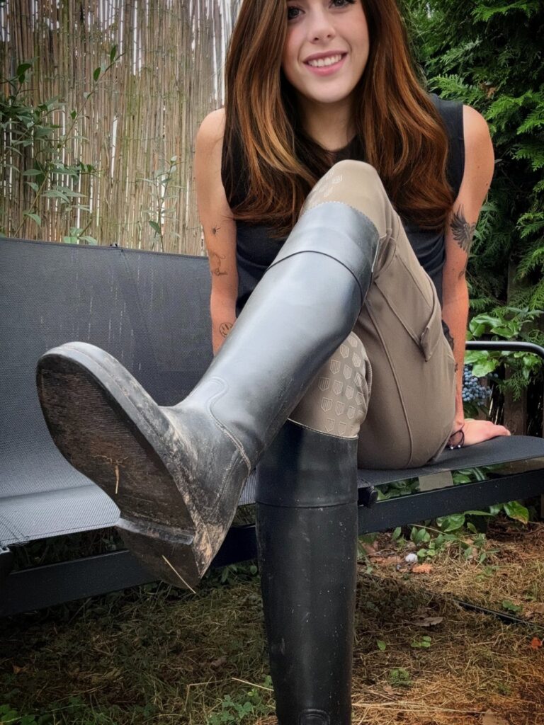 Woman in riding breeches and tall black boots sitting on a garden bench.